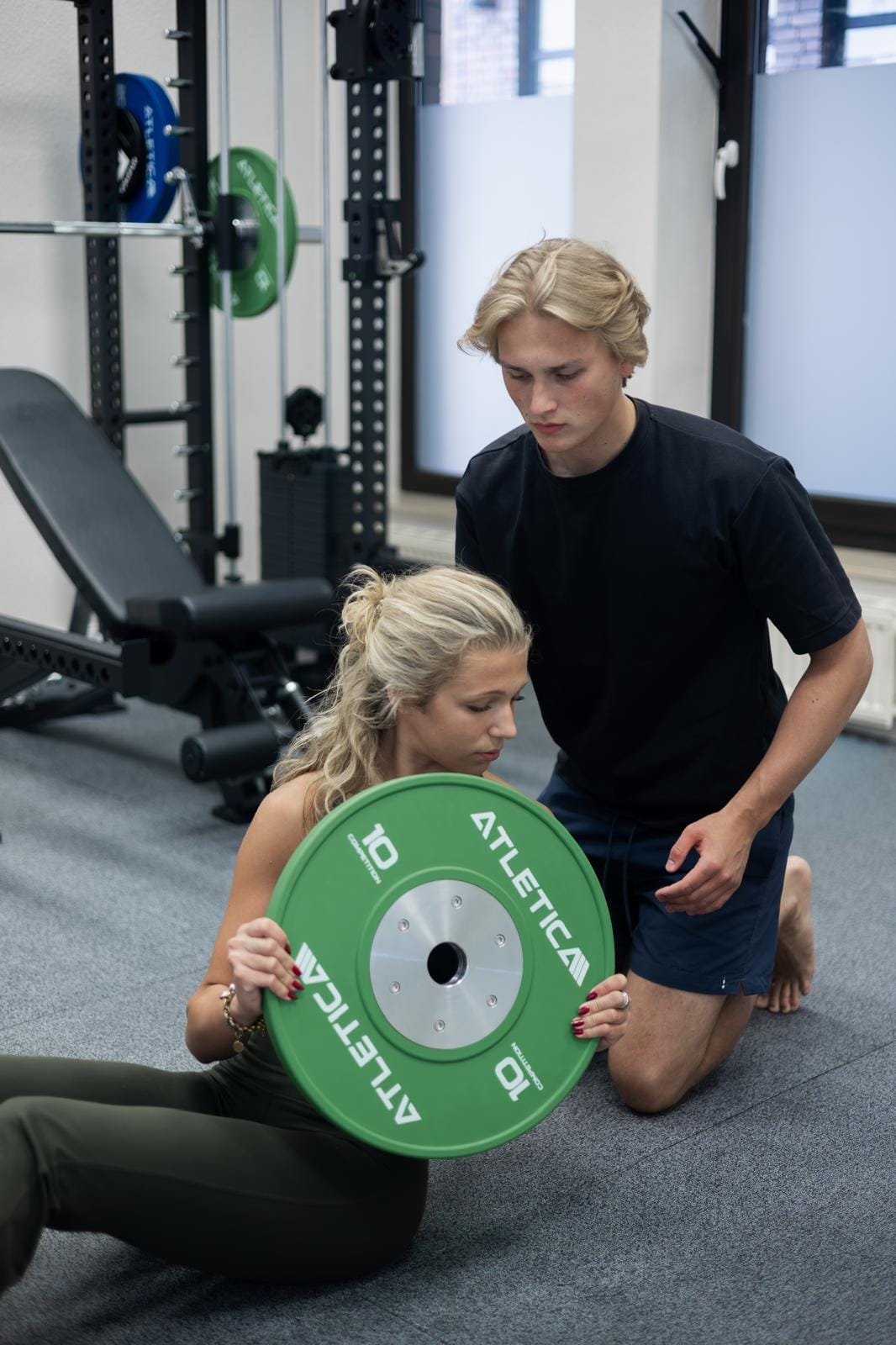 Therapeut begleitet eine Klientin bei einer Rumpfübung mit Hantelscheibe auf der Trainingsfläche